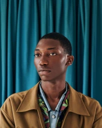 Close-up portrait of a Black man with short hair wearing a brown collar jacket over a patterned shirt against a teal curtain background