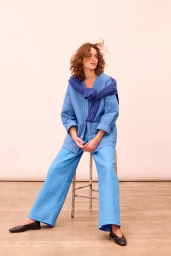 Woman with curly brown hair seated on a stool wearing a blue quilted jacket and matching blue trousers by Lucie Hugary