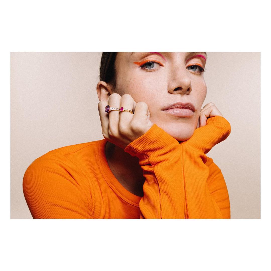 Woman with orange makeup and orange shirt poses with rings on her fingers against a light-beige background, in a beauty shot.