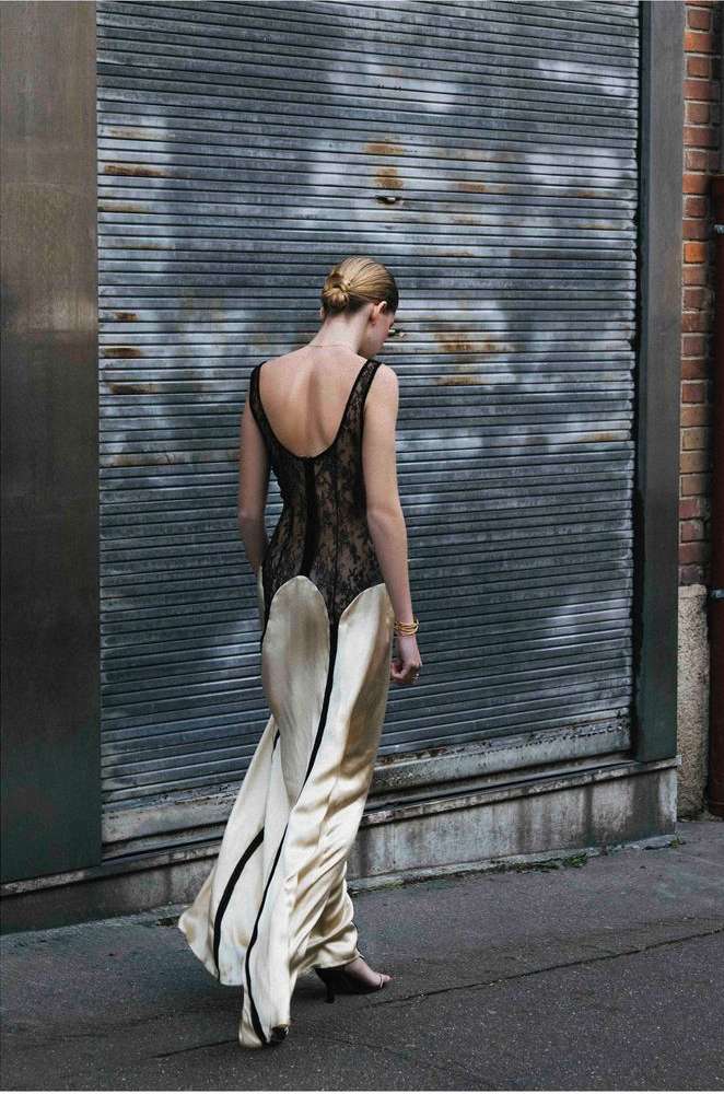 Back view of a blonde adult female model wearing a sleeveless beige and black lace gown, walking in an urban environment near a gray metal wall.