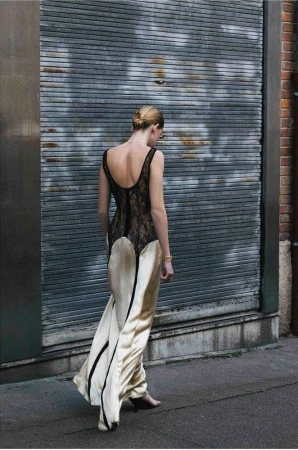 Back view of a blonde adult female model wearing a sleeveless beige and black lace gown, walking in an urban environment near a gray metal wall.