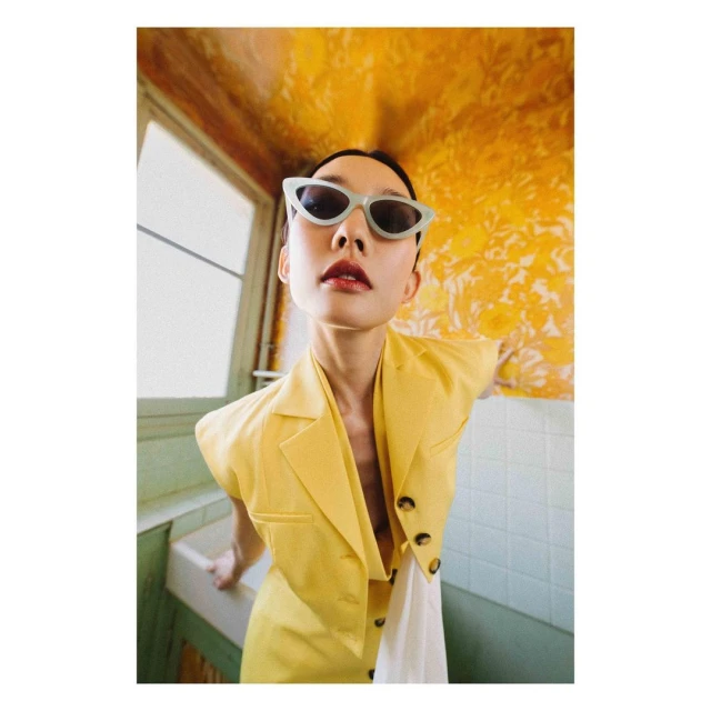 Young woman poses in a yellow blazer dress and sunglasses against a patterned yellow wall with white trim in a quirky, vintage bathroom.