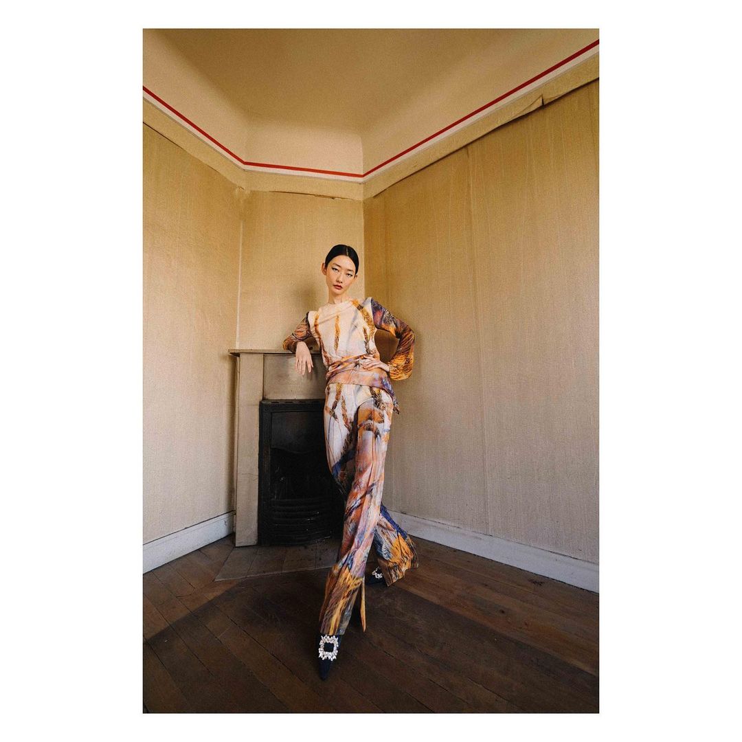 Fashion shot of a woman in a patterned jumpsuit leaning against a fireplace in an indoor setting with warm lighting and wood floors.