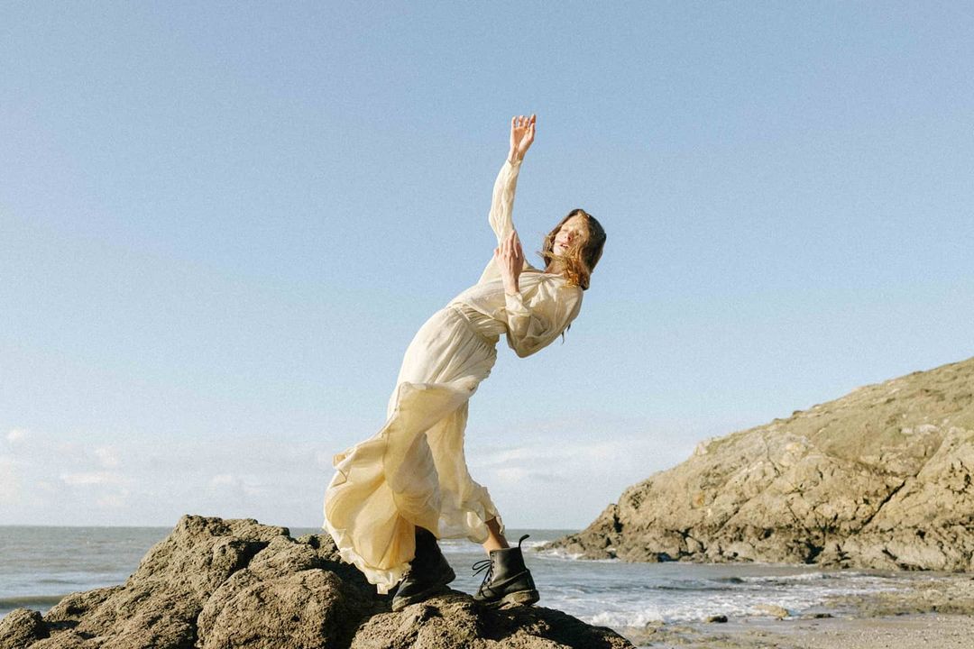 A woman in a flowing cream-colored dress and black boots poses on a rock at the beach with the ocean and a headland in the background.