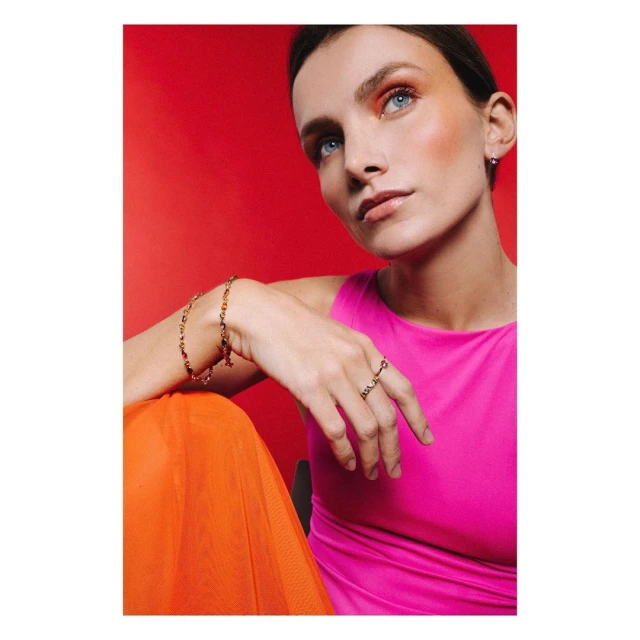 Close-up studio shot of a fair-skinned young woman with blue eyes wearing a pink top and jewelry against a bright red backdrop.
