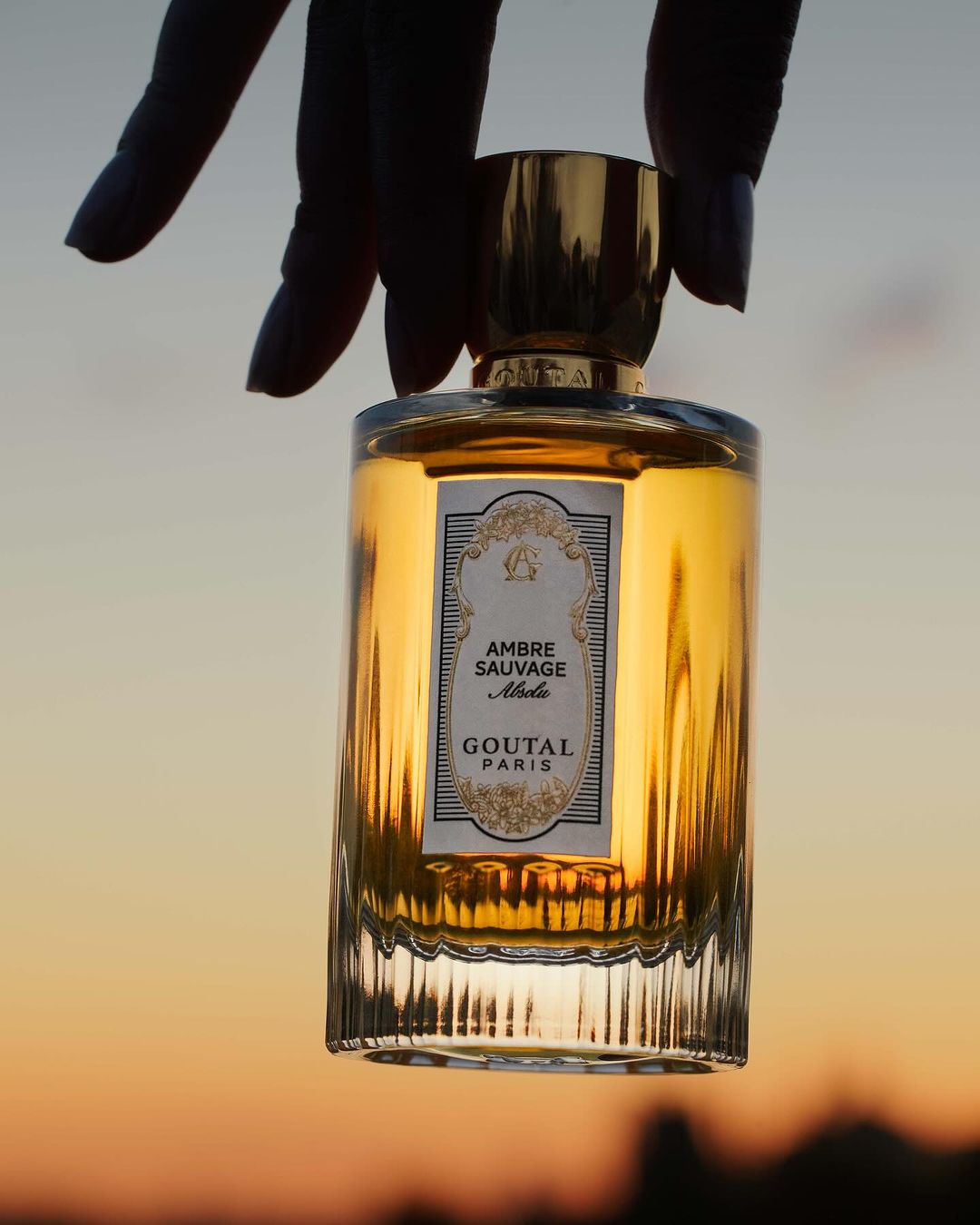 Close-up of a glass perfume bottle held in hand, featuring golden liquid and a branded label, set against a blurred sunset sky.