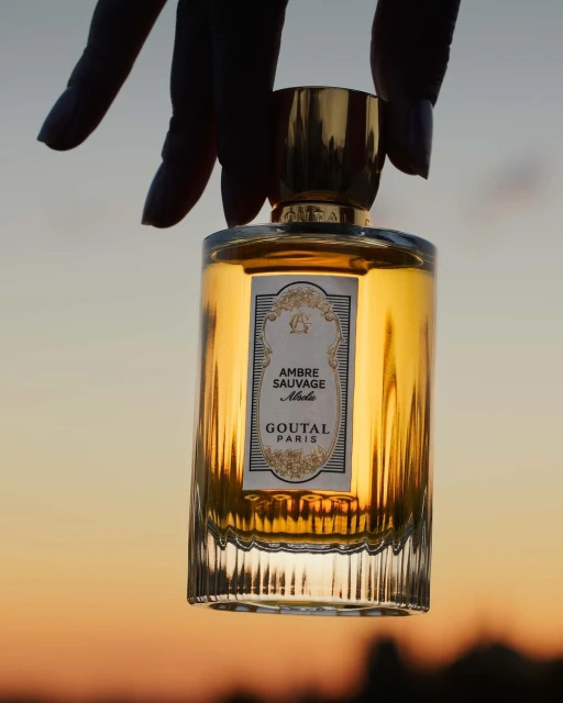Close-up of a glass perfume bottle held in hand, featuring golden liquid and a branded label, set against a blurred sunset sky.