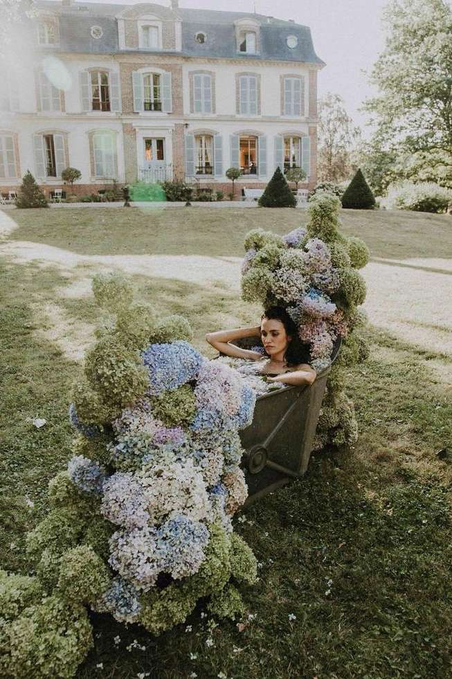 Adult female reclining in a flower-filled wheelbarrow on a lawn with a large building in the background on a sunny day.