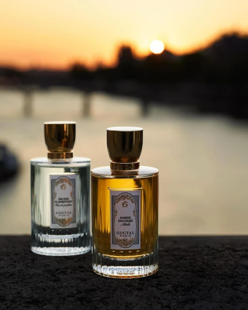 Two perfume bottles, one gold and one silver, stand against a blurred background of a bridge and a sunset sky over a river.