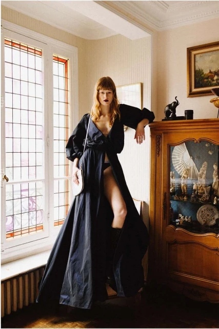 Elegant woman in a flowing black dress poses by a bright window in a classically decorated room with antique furniture and white walls.