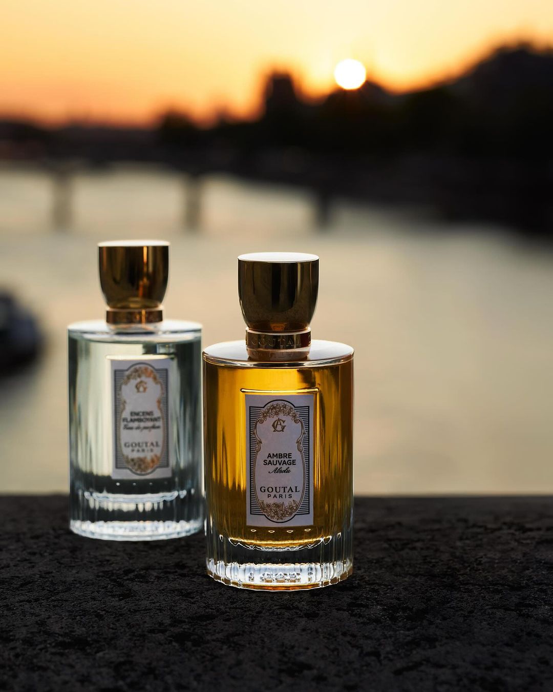Two Goutal Paris perfume bottles, one with golden liquid and one with clear liquid, stand side-by-side at sunset overlooking a river.