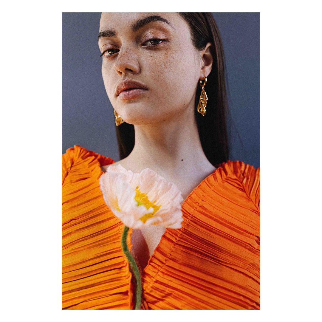 Close-up portrait of a woman wearing an orange pleated top and gold earrings, holding a pale pink flower.