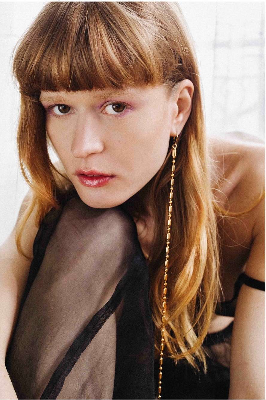 Close-up of a woman with bangs and long auburn hair wearing a long gold earring and a sheer black garment, neutral indoor background.