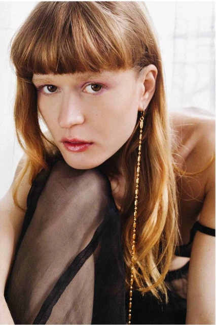 Close-up of a woman with bangs and long auburn hair wearing a long gold earring and a sheer black garment, neutral indoor background.