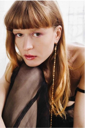 Close-up of a woman with bangs and long auburn hair wearing a long gold earring and a sheer black garment, neutral indoor background.