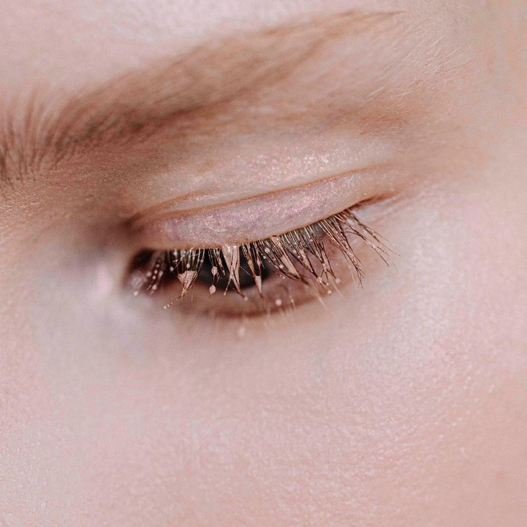 Extreme macro close-up of a closed human eye showing detailed eyelashes clustered with tiny droplets and metallic pigment.