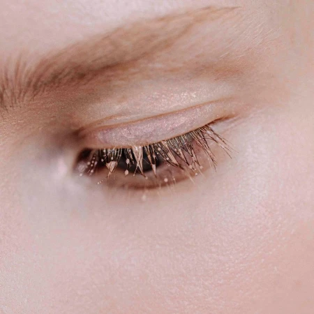 Extreme macro close-up of a closed human eye showing detailed eyelashes clustered with tiny droplets and metallic pigment.