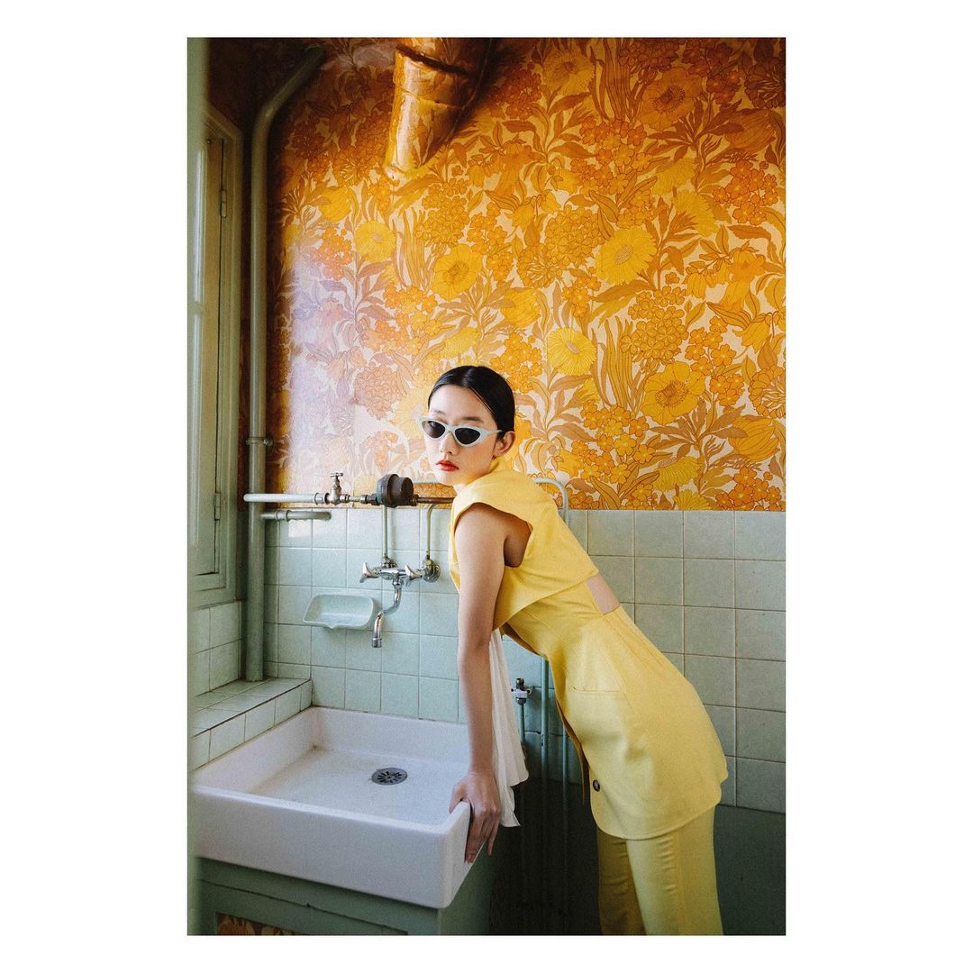 Elegant woman in yellow outfit leans on a white bathroom sink against a yellow patterned wall, wearing sunglasses, fashion photoshoot.