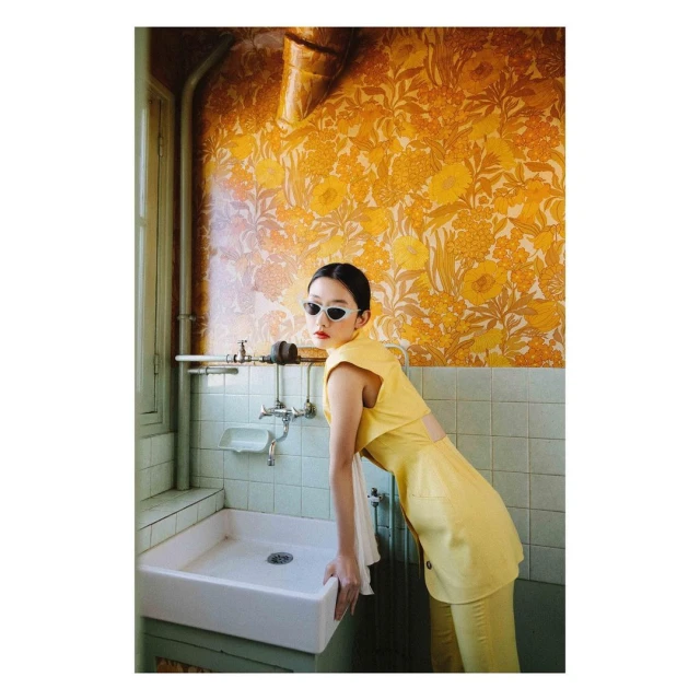 Elegant woman in yellow outfit leans on a white bathroom sink against a yellow patterned wall, wearing sunglasses, fashion photoshoot.