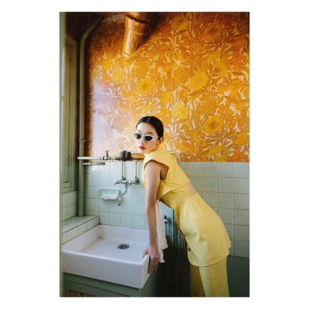 Elegant woman in yellow outfit leans on a white bathroom sink against a yellow patterned wall, wearing sunglasses, fashion photoshoot.