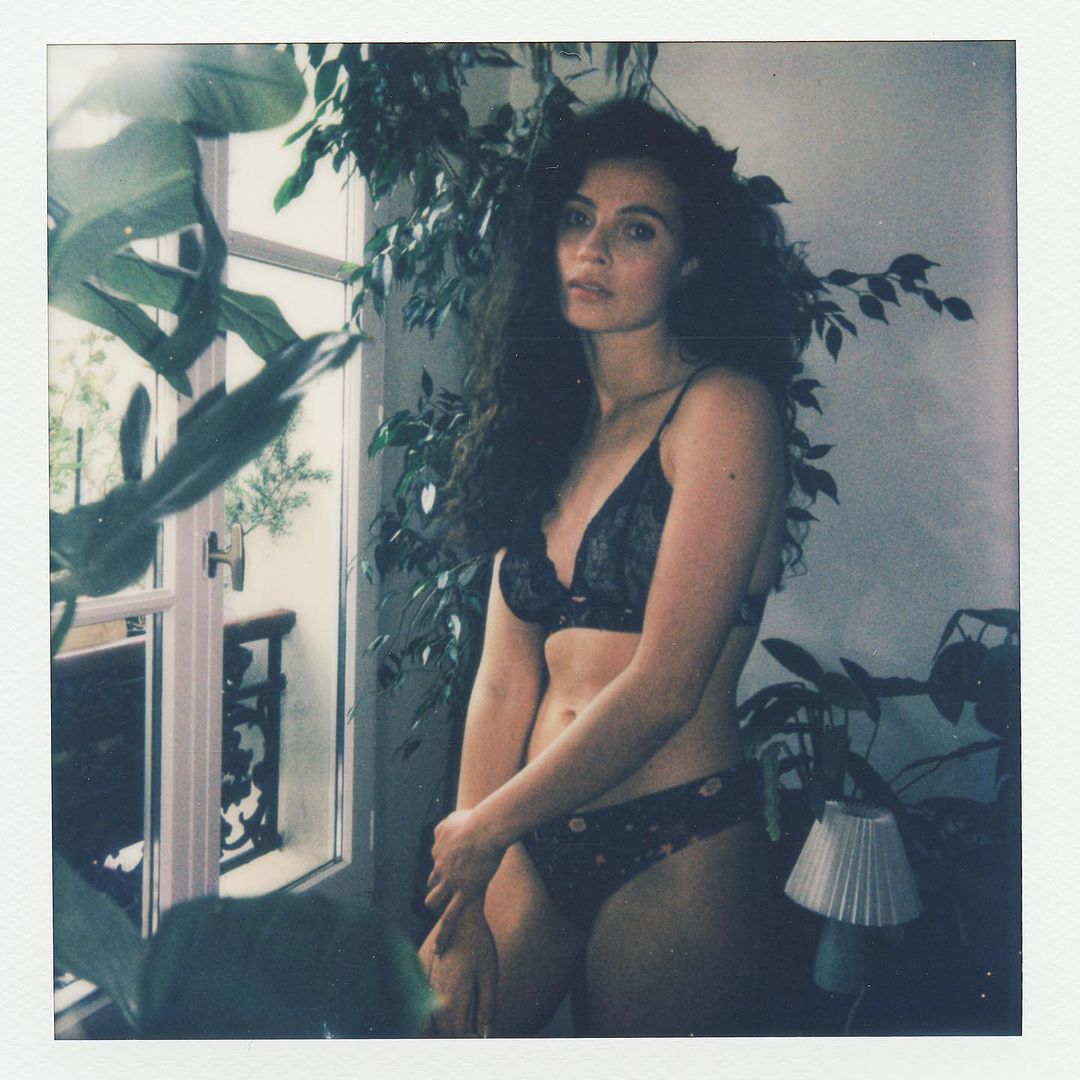 An adult female with curly hair poses in lingerie next to a window with indoor plants, creating a natural, intimate portrait.