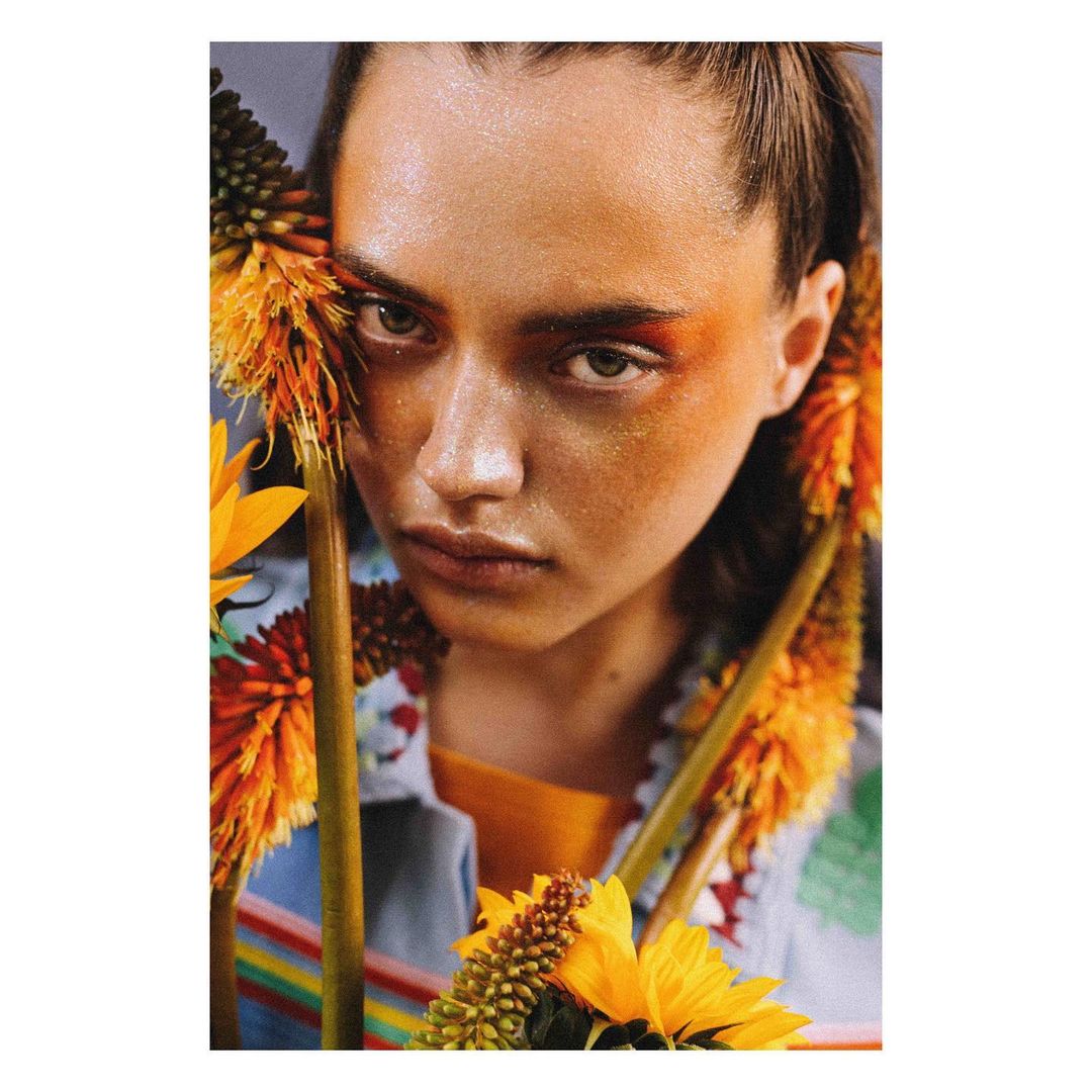 Close-up portrait of a woman with orange eyeshadow surrounded by orange and yellow flowers, wearing a colorful striped shirt.
