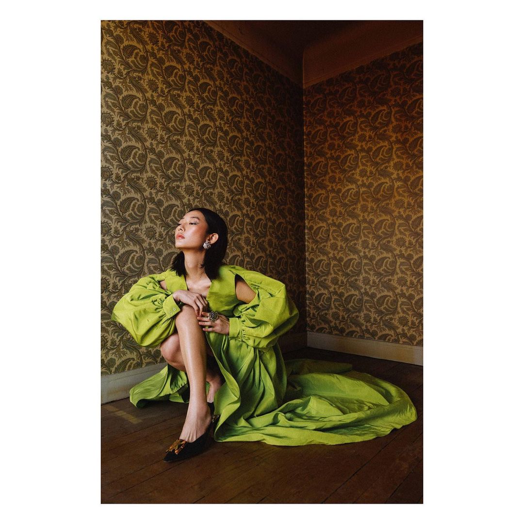 A woman in a green dress sits on a wooden floor in front of a patterned wallpaper, fashionably posed in an indoor setting.