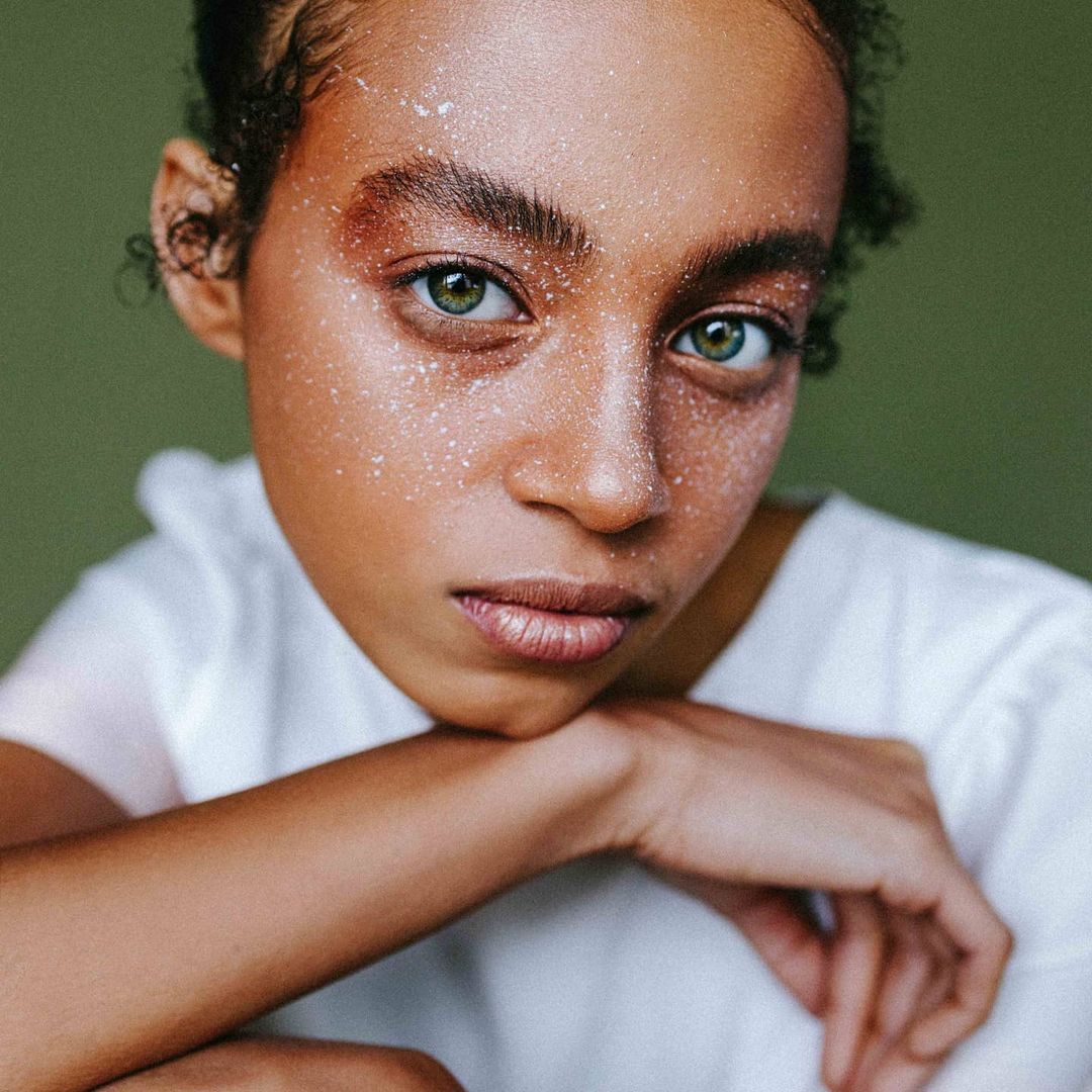Close-up portrait of a young adult woman with striking green eyes, fair skin dusted with shimmering particles, and light green background.