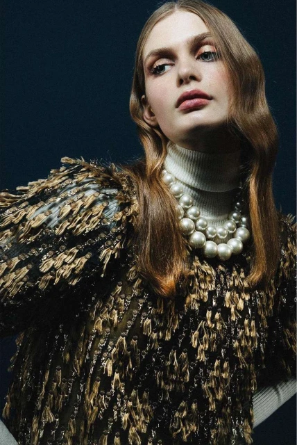 Close-up of a fair-skinned adult woman with wavy light brown hair, a white turtleneck, a gold-and-black fringed top, and a pearl necklace.