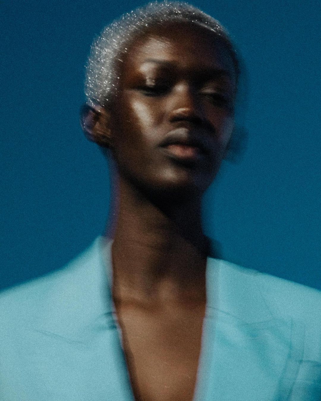 Close-up portrait of an African American adult female with metallic silver hair, wearing a blue jacket against a blue background.