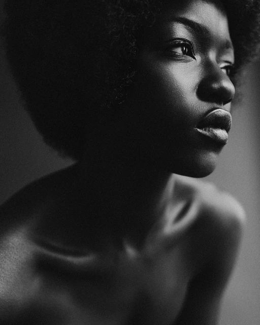 Striking black and white portrait of a Black woman with an afro looking off to the side with soft, diffused lighting in a minimalist setting.