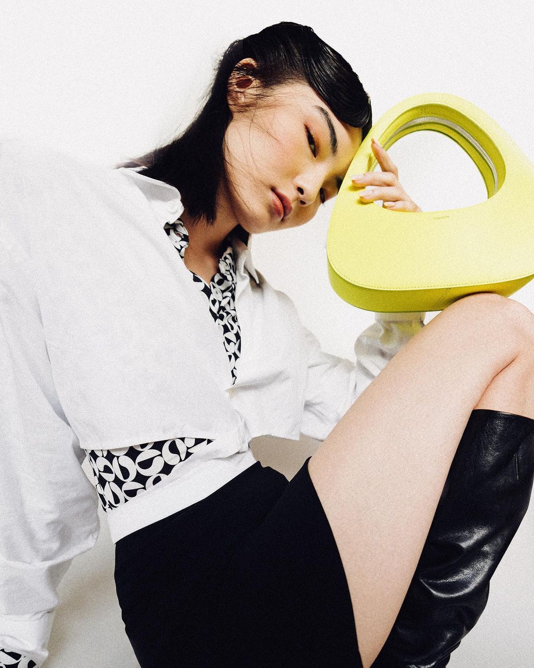 A woman poses with a bright yellow purse while wearing a white shirt, black shorts, and black boots against a white backdrop.