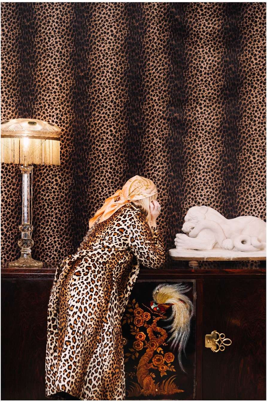 A woman in leopard print coat peers at a lion sculpture inside a room with leopard-print wallpaper, lit by a bedside lamp.