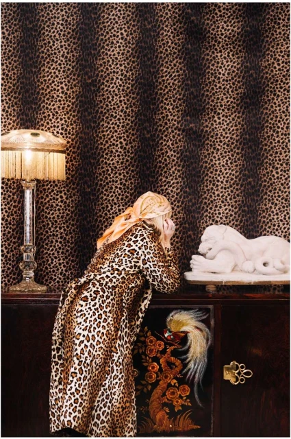 A woman in leopard print coat peers at a lion sculpture inside a room with leopard-print wallpaper, lit by a bedside lamp.