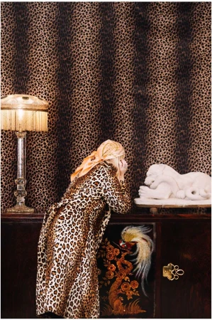 A woman in leopard print coat peers at a lion sculpture inside a room with leopard-print wallpaper, lit by a bedside lamp.