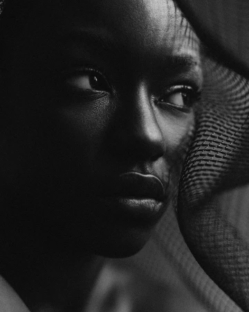 Close-up portrait of a young Black woman with dark skin, glancing off to the side, partially obscured by a dark netting.
