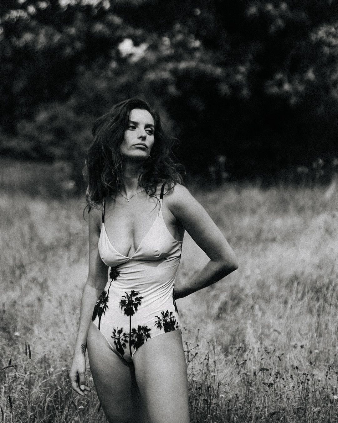 A brunette woman in a patterned swimsuit stands in a grassy field with trees in the background, posing with her hand on her hip.