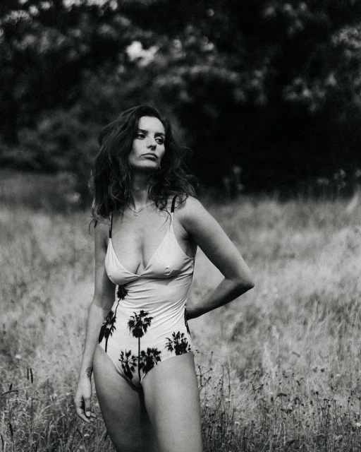 A brunette woman in a patterned swimsuit stands in a grassy field with trees in the background, posing with her hand on her hip.