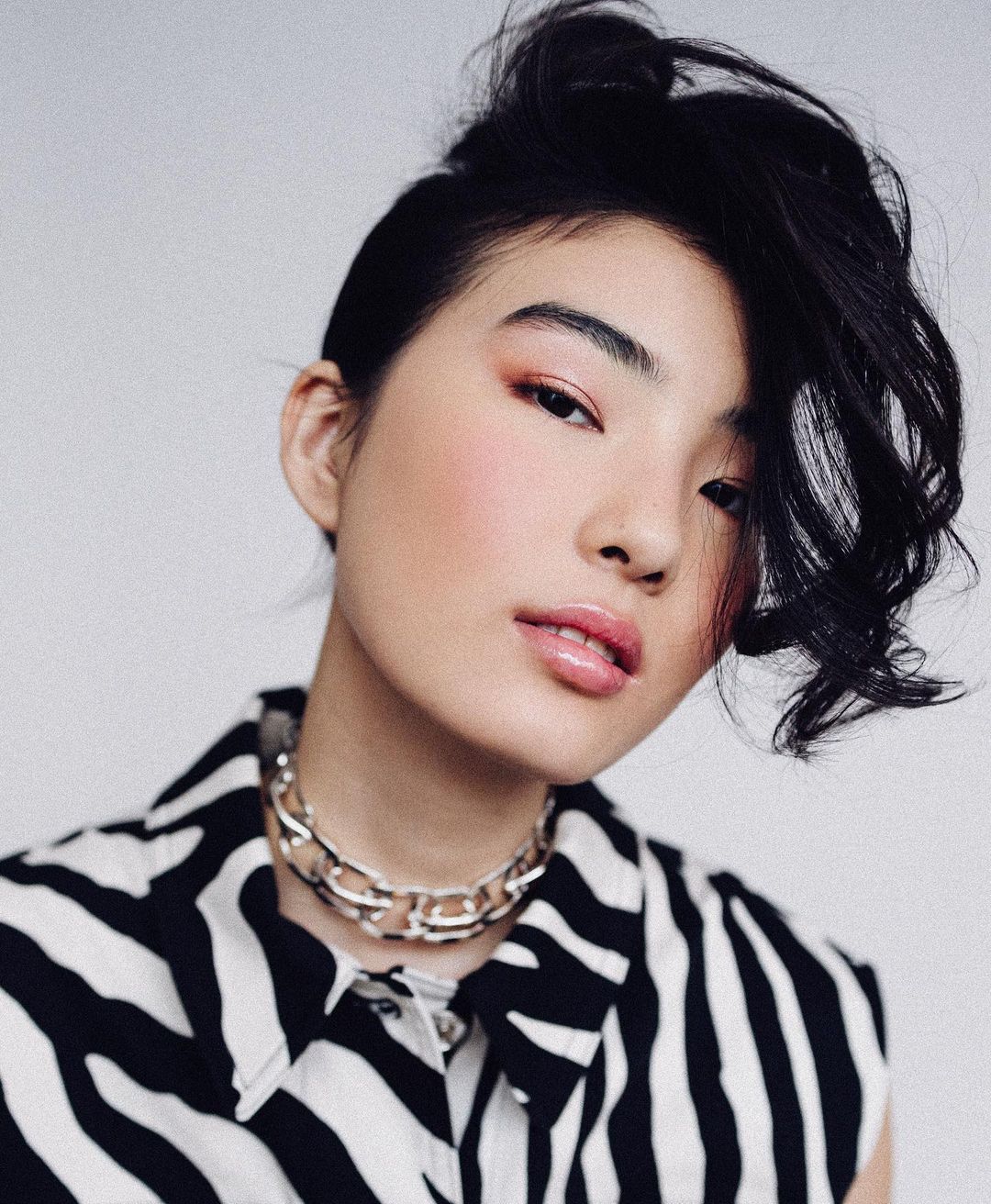 Portrait of Asian woman with black hair and zebra-print blouse against a white backdrop, wearing silver choker necklace and pink blush.