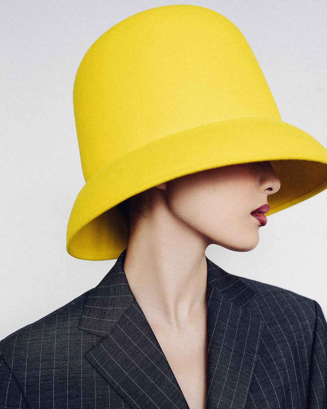 Profile view of a person wearing a yellow bucket hat and a gray pinstripe blazer against a pale background in a fashion-style photograph.