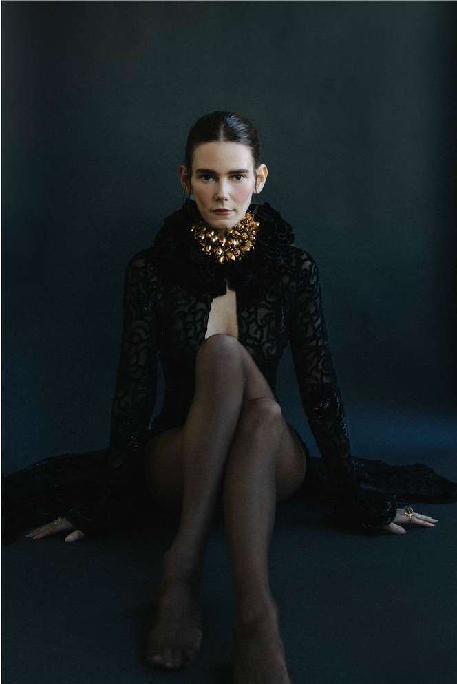 Portrait of a woman with dark hair, gold necklace, sheer black robe, and dark tights, sitting cross-legged against a dark backdrop.