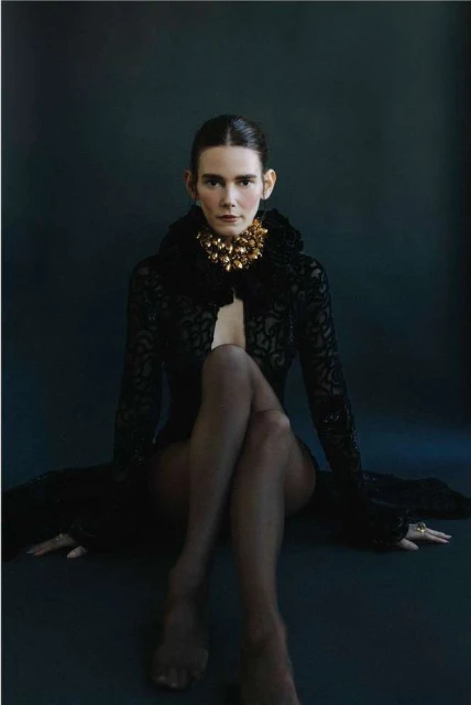 Portrait of a woman with dark hair, gold necklace, sheer black robe, and dark tights, sitting cross-legged against a dark backdrop.
