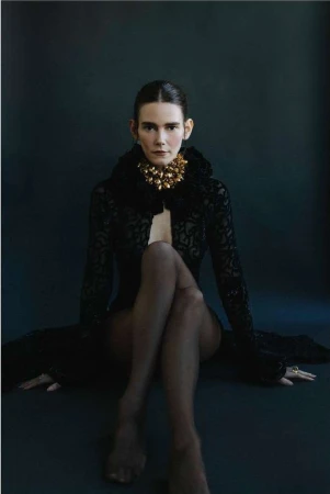 Portrait of a woman with dark hair, gold necklace, sheer black robe, and dark tights, sitting cross-legged against a dark backdrop.