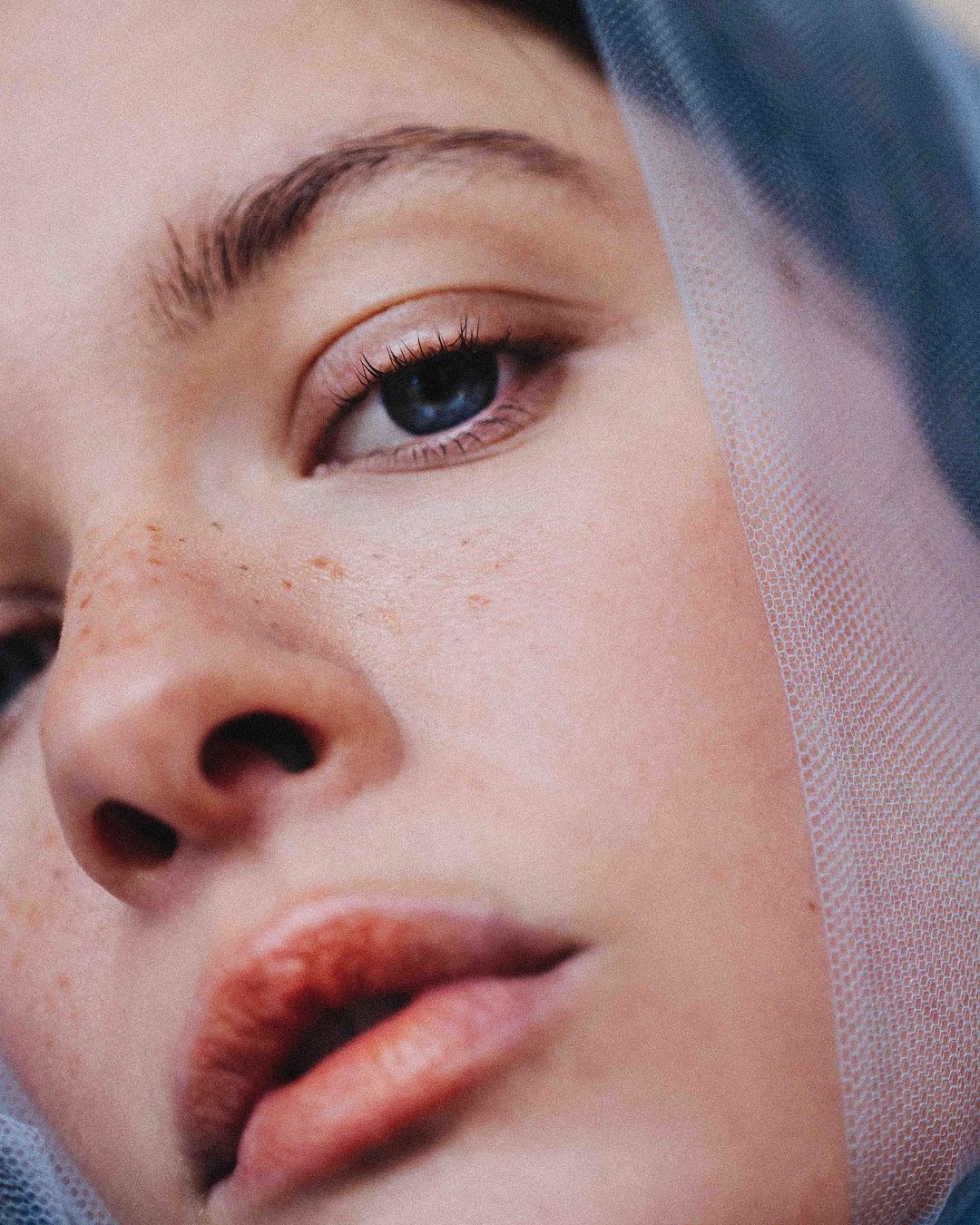 Close-up portrait of an adult female with blue eyes and freckled skin, partially covered by a sheer blue veil in soft, diffused light.