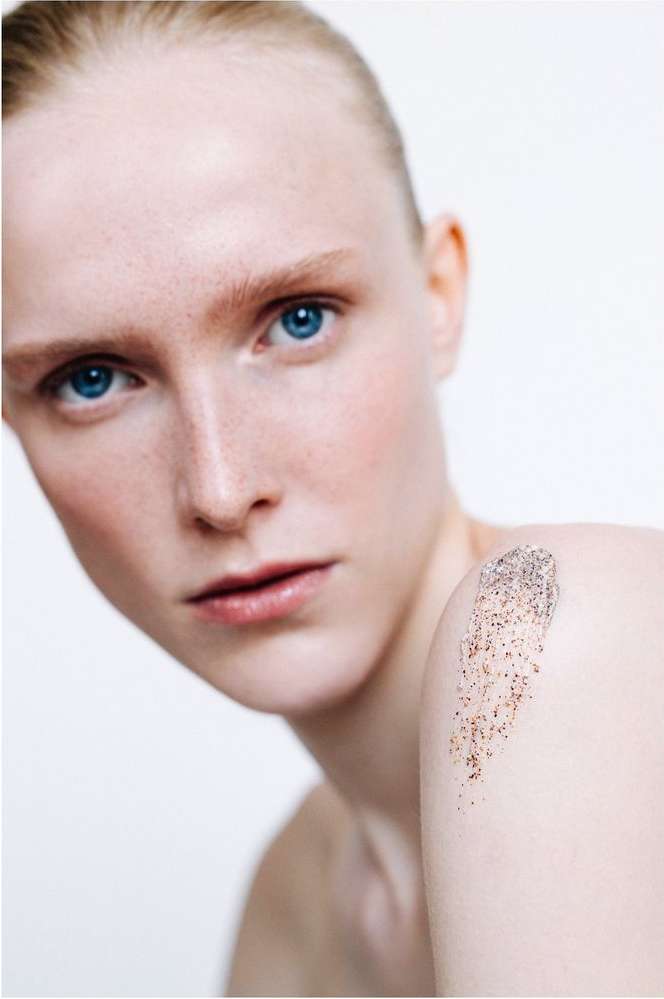 Close-up of a fair-skinned woman with blue eyes and freckles, fair hair, and facial scrub on shoulder, on a white background.