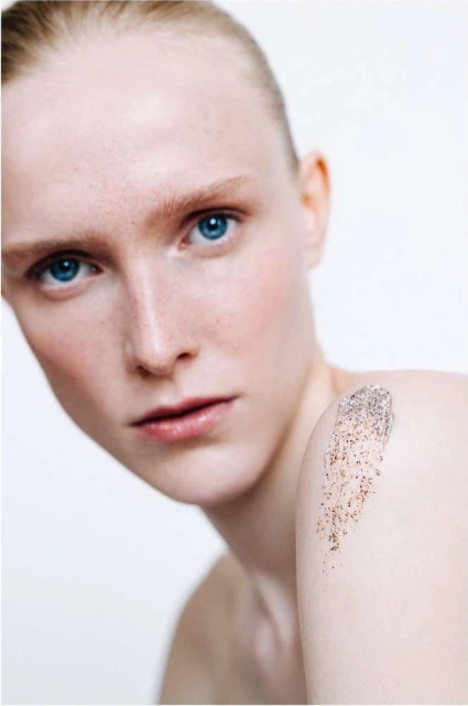 Close-up of a fair-skinned woman with blue eyes and freckles, fair hair, and facial scrub on shoulder, on a white background.
