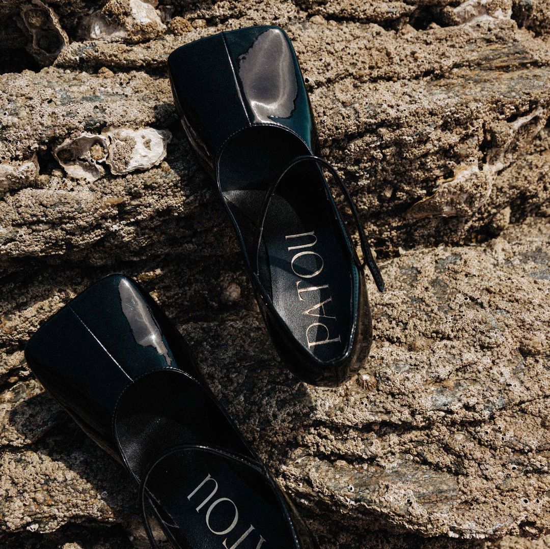 Close-up of two glossy black Patou ballet flats with ankle straps resting on a textured, sandy-colored rock surface in natural light.