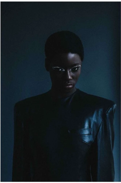 Close-up portrait of a dark-skinned woman with white eyeliner, wearing a black jacket, against a dark blue background in a studio setting.