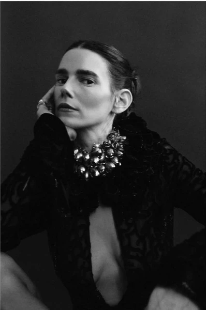 Portrait of adult woman with hand near her face. The woman is wearing a statement necklace and a patterned, low-cut top.