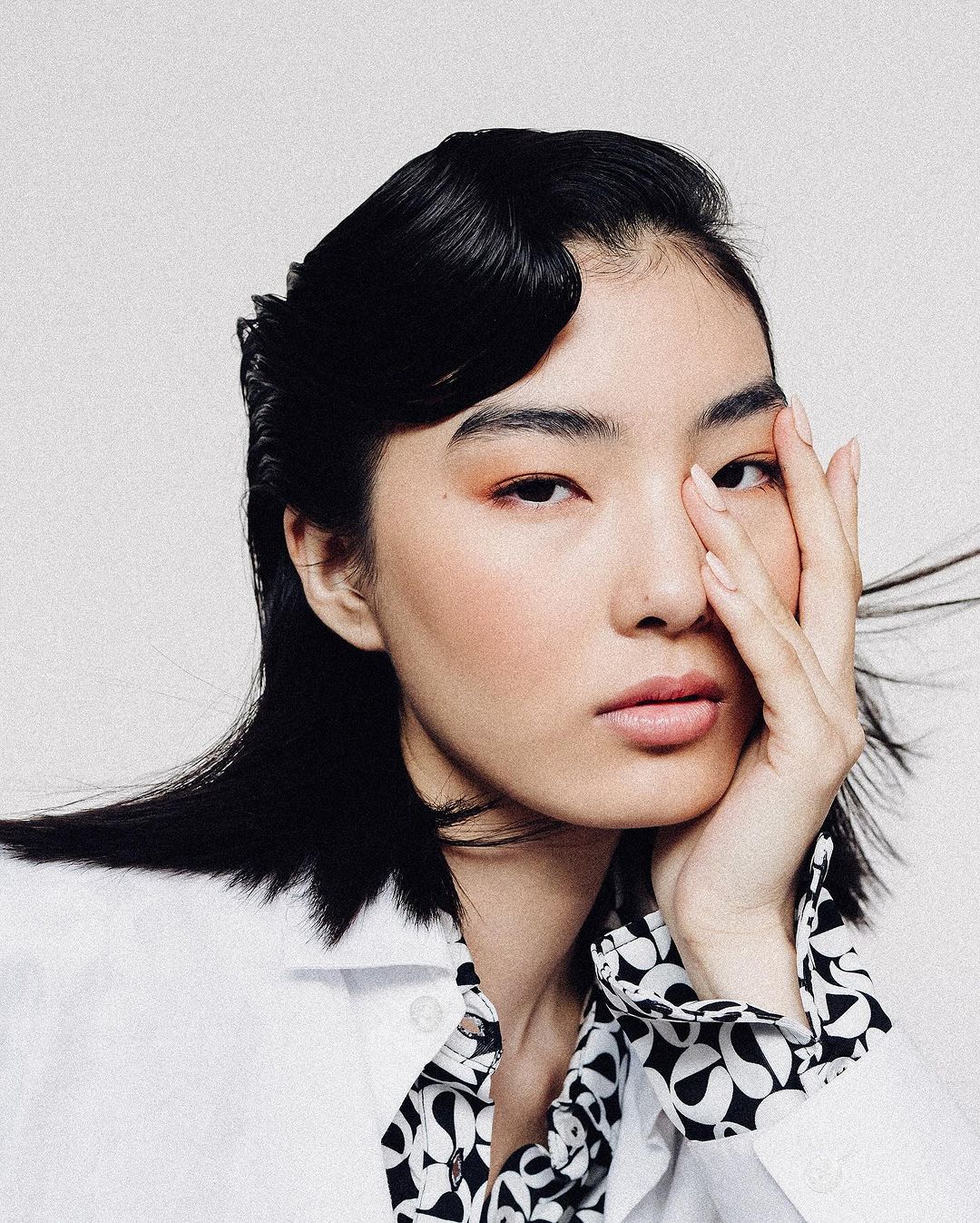 Portrait of a woman with black hair and hand near face wearing a patterned shirt against a white backdrop